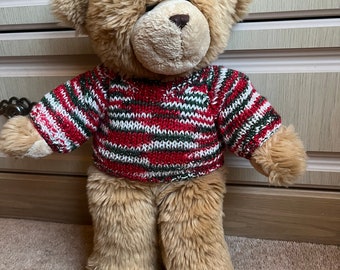 Può includere: Un orsacchiotto marrone che indossa un maglione a righe rosse, verdi e bianche. Il maglione ha una finitura scintillante.