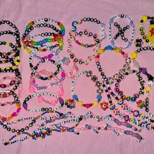 Könnte beinhalten: Eine Sammlung bunter Perlenarmbänder mit verschiedenen Phrasen und Designs. Die Armbänder haben Buchstabenperlen mit Wörtern wie "GAY", "BE KIND" und "TRANS RIGHTS". Einige Armbänder haben Stern-, Herz- und Blumenakzente. Der Hintergrund ist ein hellrosa Stoff.