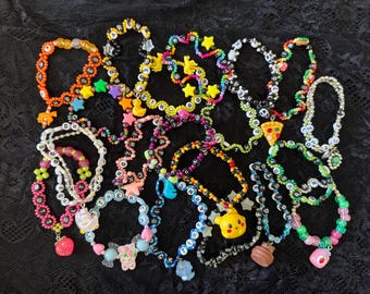 Könnte beinhalten: Eine Sammlung farbenfroher Perlenketten und Armbänder. Der Schmuck verfügt über verschiedene Anhänger, darunter Sterne, Enten und Pizzastücke. Die Perlen sind in einer Vielzahl von Farben gehalten, darunter Orange, Gelb, Rosa und Blau. Einige Teile haben Buchstabenperlen.