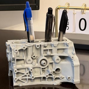 May include: A gray, 3D printed engine block pen holder with three pens inserted. The pens are blue, black, and black. The pen holder is sitting on a dark brown wooden surface. A gold calendar with white cards displaying the numbers "02" is in the background.