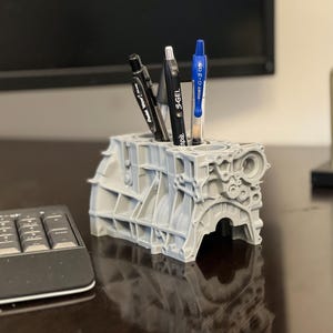Może przedstawiać: Szary, 3D-drukowany stojak na długopisy w kształcie bloku silnika z trzema długopisami w środku. Długopisy są czarne i niebieskie. Stojak na długopisy stoi na biurku z klawiaturą i kalendarzem.