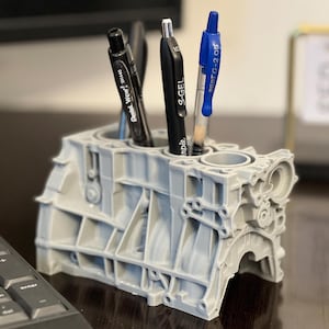 Może przedstawiać: Szary, 3D-wydrukowany stojak na długopisy w kształcie bloku silnika z czterema długopisami w środku. Długopisy są czarne, czarne i srebrne, czarne i srebrne oraz niebieskie. Stojak na długopisy stoi na ciemnym drewnianym blacie.