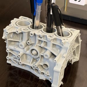 Może przedstawiać: Szary organizer na biurko w kształcie bloku silnika, wydrukowany w 3D, z trzema długopisami w środku. Długopisy są niebieskie, czarne i przezroczyste.