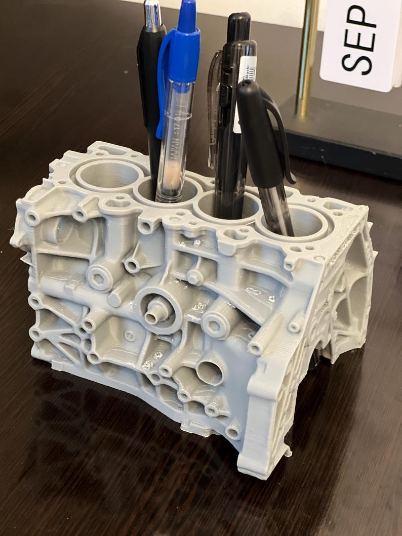 Może przedstawiać: Szary organizer na biurko w kształcie bloku silnika, wydrukowany w 3D, z trzema długopisami w środku. Długopisy są niebieskie, czarne i przezroczyste.