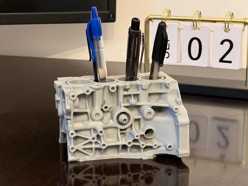 May include: A gray, 3D printed engine block pen holder with three pens inserted. The pens are blue, black, and black. The pen holder is sitting on a dark brown wooden surface. A gold calendar with white cards displaying the numbers "02" is in the background.