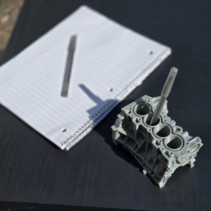 Może przedstawiać: Szary, tr&oacute;jwymiarowy model bloku silnika z czterema cylindrami, wydrukowany w technologii 3D. Model służy jako stojak na długopisy, w kt&oacute;ry włożono srebrny długopis do jednego z cylindr&oacute;w.