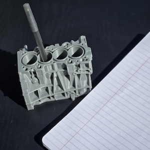 Może przedstawiać: Szary, tr&oacute;jwymiarowy model bloku silnika z czterema cylindrami, używany jako stojak na długopisy. Czarny długopis jest włożony do jednego z cylindr&oacute;w.