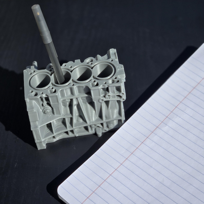 Może przedstawiać: Szary, tr&oacute;jwymiarowy model bloku silnika z czterema cylindrami, używany jako stojak na długopisy. Czarny długopis jest włożony do jednego z cylindr&oacute;w.