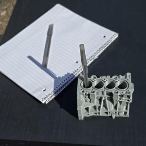 Może przedstawiać: Szary, tr&oacute;jcylindrowy blok silnika wydrukowany w 3D. Dwa długopisy umieszczone są w cylindrach. Blok silnika znajduje się na czarnej powierzchni.