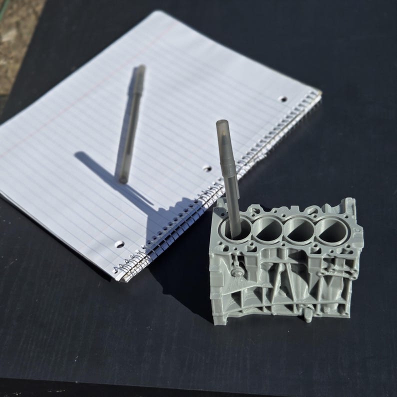 Może przedstawiać: Szary, tr&oacute;jcylindrowy blok silnika wydrukowany w 3D. Dwa długopisy umieszczone są w cylindrach. Blok silnika znajduje się na czarnej powierzchni.