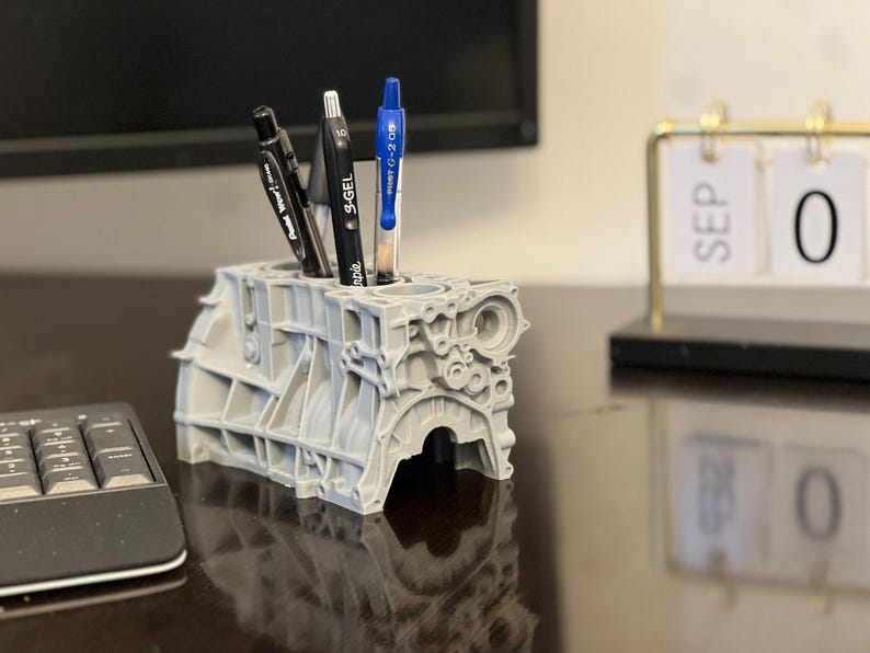Może przedstawiać: Szary, 3D-drukowany stojak na długopisy w kształcie bloku silnika z trzema długopisami w środku. Długopisy są czarne i niebieskie. Stojak na długopisy stoi na biurku z klawiaturą i kalendarzem.