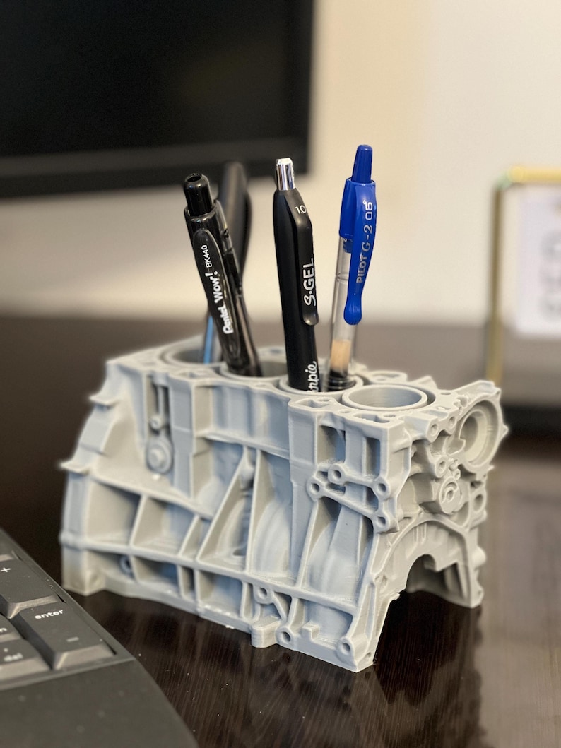 Może przedstawiać: Szary, 3D-wydrukowany stojak na długopisy w kształcie bloku silnika z czterema długopisami w środku. Długopisy są czarne, czarne i srebrne, czarne i srebrne oraz niebieskie. Stojak na długopisy stoi na ciemnym drewnianym blacie.