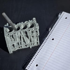 Może przedstawiać: Szary, tr&oacute;jwymiarowy model bloku silnika z czterema cylindrami, używany jako organizer na biurko. Pi&oacute;ro jest włożone do jednego z cylindr&oacute;w.