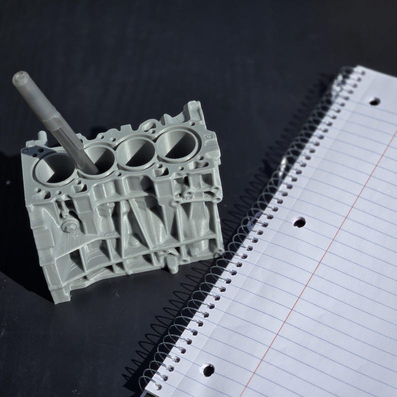 Może przedstawiać: Szary, tr&oacute;jwymiarowy model bloku silnika z czterema cylindrami, używany jako organizer na biurko. Pi&oacute;ro jest włożone do jednego z cylindr&oacute;w.