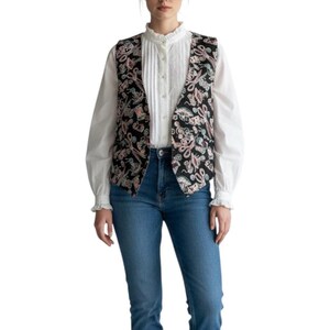 May include: A black vest featuring a colorful pattern of sewing tools including scissors, spools of thread, and needles. The vest is worn over a white long-sleeved blouse with a high ruffled collar and vertical pleats, paired with blue denim jeans.