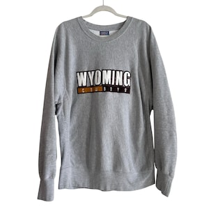 Puede incluir: Una sudadera gris jaspeada con la palabra "WYOMING" en letras blancas sobre "COWBOYS" en marrón y blanco. La sudadera tiene cuello redondo y mangas largas.