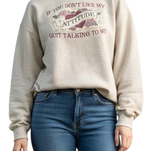 May include: Beige sweatshirt with the text "IF YOU DON'T LIKE MY ATTITUDE QUIT TALKING TO ME!" in a decorative design. The sweatshirt is paired with blue jeans. The design features a heart and floral elements.
