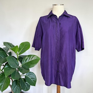 以下が含まれることがあります： 鮮やかな紫色のボタンダウンシャツ、半袖。白い背景にマネキンが飾られており、左側には緑の葉の植物があります。シャツはクラシックな襟と、少しオーバーサイズのフィット感です。