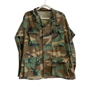 May include: A long-sleeve camouflage jacket featuring a woodland pattern in shades of green, brown, and black. The jacket includes a collar, a button-front closure, two chest pockets with flaps, and two lower pockets. A U.S. ARMY patch is on the chest.