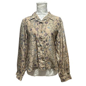 Puede incluir: Una camisa de manga larga con botones en color tostado claro que presenta un patrón repetitivo de enredaderas florales en violeta oscuro y azul claro. La camisa tiene un cuello clásico, tapeta frontal con botones y puños abotonados en un maniquí.