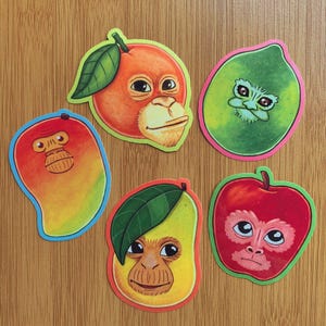 May include: Six colourful fruit stickers, each featuring a monkey-like face. The stickers depict an orange, a lime, a red apple, and two mangoes. Each sticker has a unique design and a playful expression.