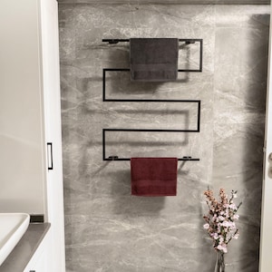 May include: A black heated towel rack with two towels, one dark gray and one burgundy, hanging on it. The rack is mounted on a gray marble wall in a bathroom.