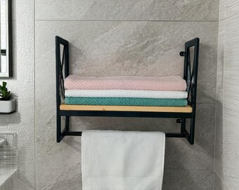 May include: A black metal and wood bathroom shelf with a towel rack. The shelf holds a stack of folded towels in pink, white, and teal. A white towel hangs from the rack below. The shelf is mounted on a gray tiled wall.