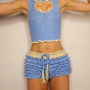 May include: A blue crocheted crop top with a heart-shaped cutout and a white crocheted trim. The top is paired with matching blue crocheted shorts with a ruffled hem and a white drawstring.