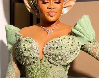 Op de afbeelding: Een vrouw die een lichtgroene, met kralen bezette jurk draagt met een bijpassende hoofddoek. De jurk heeft een diepe V-hals en lange mouwen. De hoofddoek is bovenop haar hoofd in een knoop geknoopt. De vrouw kijkt naar beneden en haar ogen zijn gesloten.