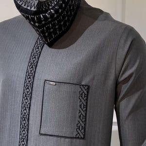 May include: A grey traditional outfit, comprising a long-sleeved shirt with a pocket and decorative trim, and a matching cap. The cap features a black and white geometric pattern. The outfit is made of a textured fabric.