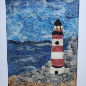 May include: A needle felted artwork depicting a red and white striped lighthouse on a rocky shore with blue water and a cloudy sky.