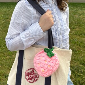 May include: A white canvas tote bag with blue handles and a pink crocheted strawberry charm. The bag has the Trader Joe's logo printed on the front.