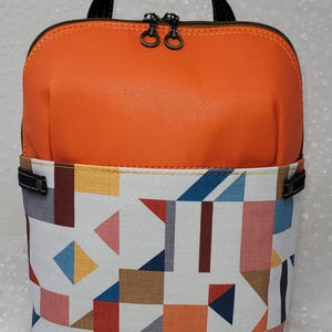 Könnte beinhalten: Orangefarbener Rucksack mit geometrischem Muster in Weiß, schwarzen Trägern und Reißverschluss.