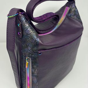Puede incluir: Un bolso de hombro de cuero morado con un diseño estampado floral y detalles de cremallera iridiscente.