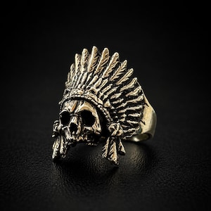 May include: A metallic gold-toned ring featuring a detailed human skull wearing a large, ornate feathered headdress. The design includes deep, dark recessed areas that highlight the intricate textures of the feathers and the skull's facial features.