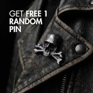 Può includere: Una spilla in metallo color argento a forma di teschio con ossa incrociate fissata su una giacca di pelle scura e usurata. Il testo GET FREE 1 RANDOM PIN è stampato in bianco nell'angolo in alto a sinistra, in contrasto con lo sfondo.