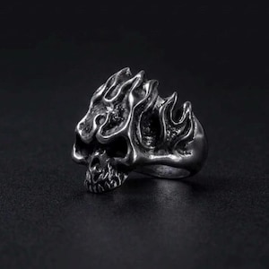 May include: A silver-toned metal ring featuring a detailed skull design integrated with flame-like patterns rising from the top of the cranium. The surface has a dark, oxidized finish in the recessed areas to highlight the sculpted texture.
