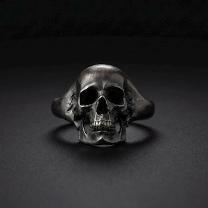Könnte beinhalten: Ein Ring aus dunkel oxidiertem Silber mit einem detaillierten, realistischen menschlichen Totenkopf-Design. Der Schädel hat tiefe, hohle Augenhöhlen, eine definierte Nasenhöhle und sichtbare Zähne, wobei die Ringschiene nahtlos in die Seiten übergeht.