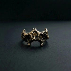 May include: A bronze ring with a distinctive, textured design. The ring has an openwork centre with a small, circular accent. The overall style is rustic and artistic, set against a dark background.