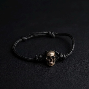 Puede incluir: Una pulsera de cordón negro con una cuenta central de calavera metálica con acabado en bronce desgastado. El cordón está asegurado con nudos ajustables a ambos lados de la calavera, creando un diseño de banda circular con textura oscura.