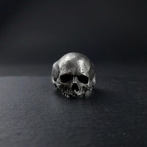 Può includere: Un anello a forma di teschio argentato e dettagliato. L'anello presenta un design realistico a forma di teschio con orbite vuote e una mascella dettagliata. Lo sfondo scuro mette in risalto i dettagli intricati e l'estetica gotica.