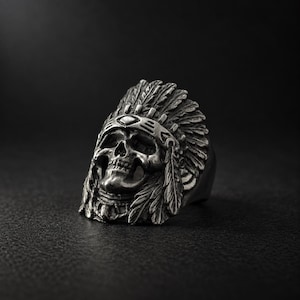 May include: A silver-toned metal ring featuring a detailed skull wearing a traditional feathered headdress. The skull has deep eye sockets and a nasal cavity, while the headdress displays intricate carved feather textures and a central band.