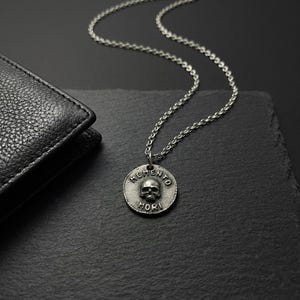Puede incluir: Collar con colgante plateado con diseño de calavera. El colgante redondo tiene las palabras "MEMENTO MORI" alrededor de la calavera. El collar está sobre una superficie negra junto a una cartera de cuero negra.