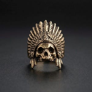 Puede incluir: Un anillo detallado de color bronce que presenta una calavera con un tocado de plumas. El anillo tiene detalles intrincados en la calavera y las plumas, creando un accesorio único y audaz.