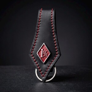 Puede incluir: Un llavero de cuero negro con un emblema metálico en forma de diamante que presenta un fondo rojo texturizado y el texto plateado 1%. El cuero está cosido con hilo rojo en los bordes e incluye una anilla de metal plateado en la base.