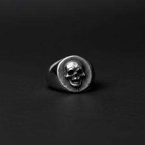 May include: A silver signet ring with a detailed skull design. The skull is centered on a circular face, with a polished band. The ring is set against a dark background, highlighting the metallic sheen and intricate details of the skull.