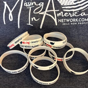May include: White silicone bracelets with the text "Missing in America Network.com" and "You are never alone!" printed on them. The bracelets are on a grey shirt with the text "Missing in America Network.com 501c3 Non Profit" printed on it.