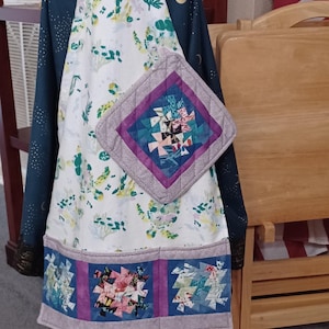 Puede incluir: Un delantal blanco con estampado floral y un agarrador de olla acolchado morado y azul. El delantal tiene tres bolsillos con cuadrados acolchados en la misma tela que el agarrador de olla.