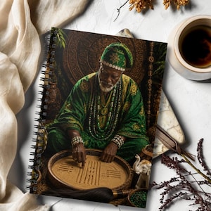 May include: A spiral-bound notebook featuring an illustration of an elderly person wearing a green patterned robe, matching cap, and beaded necklaces. The person is seated, pointing at a circular sand tray with markings, surrounded by decorative items.