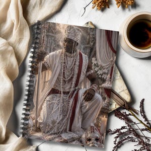 May include: A spiral-bound notebook featuring a printed image of a person in white and red ceremonial attire, wearing a crown and multiple beaded necklaces, kneeling while holding a long sword. The notebook is displayed with a cup of coffee.